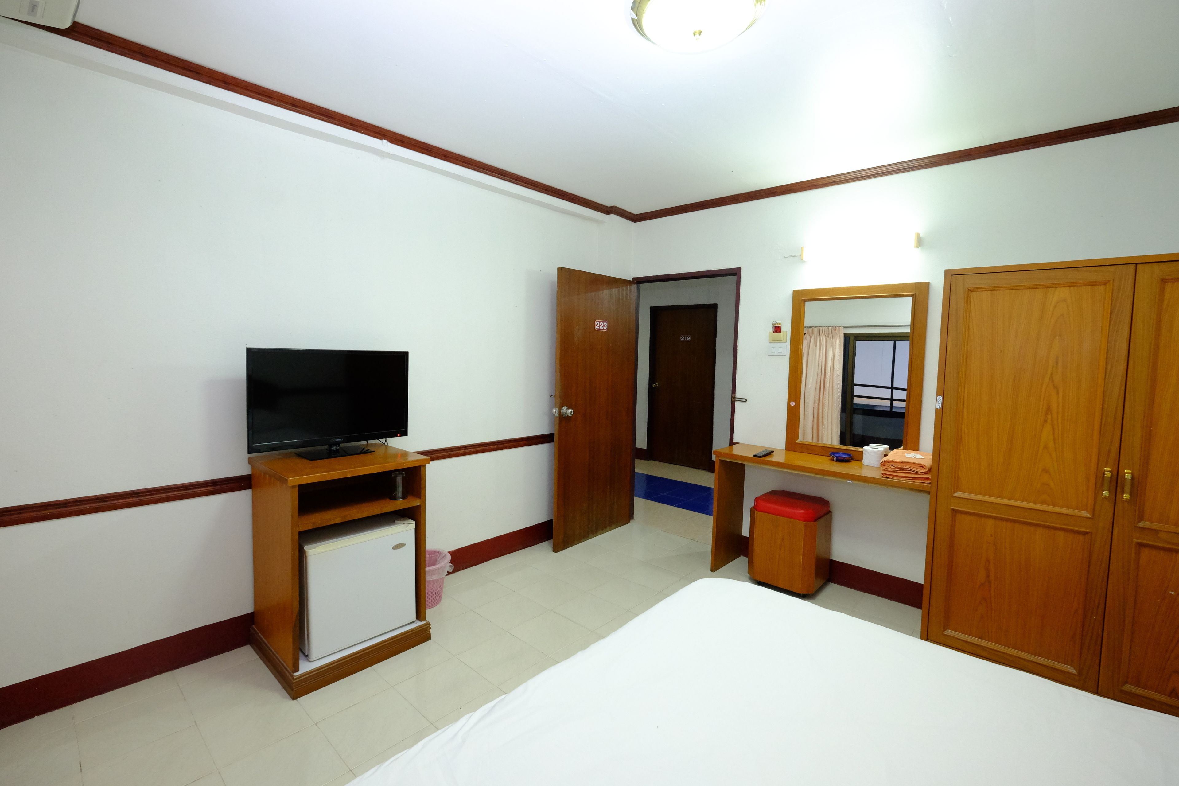 A hotel room with a bed, a television on a wooden stand, a small refrigerator, a wooden wardrobe, a mirror, and a door leading to another room.