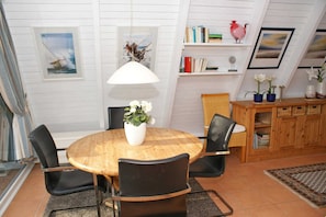 Un coin repas avec une table ronde en bois, des chaises noires, un vase blanc orné de fleurs et une suspension blanche. On y trouve également des étagères remplies de livres et d’objets décoratifs, ainsi qu’un meuble en bois contenant des paniers.