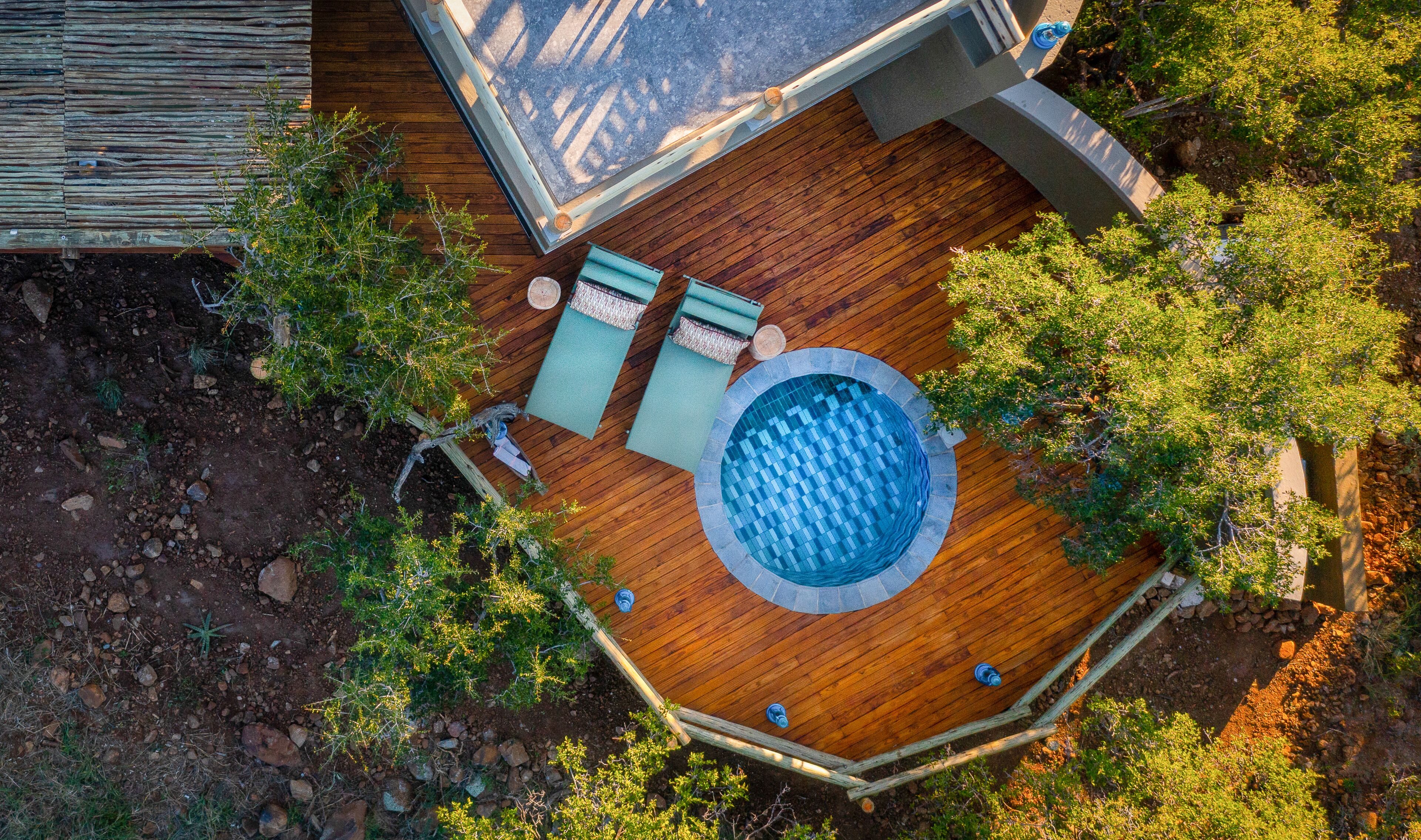 An aerial view of a wooden deck with a circular pool, surrounded by trees and a stone wall.