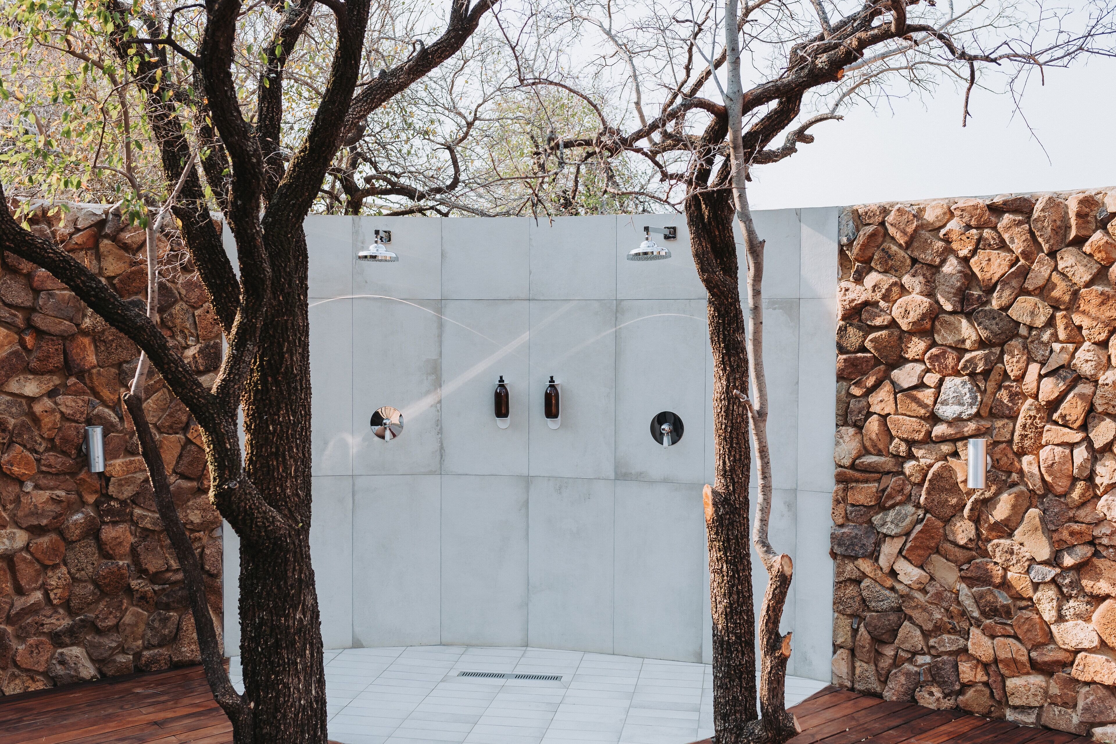 Zona de ducha al aire libre con paredes de piedra, suelo de baldosas y rodeada de árboles.