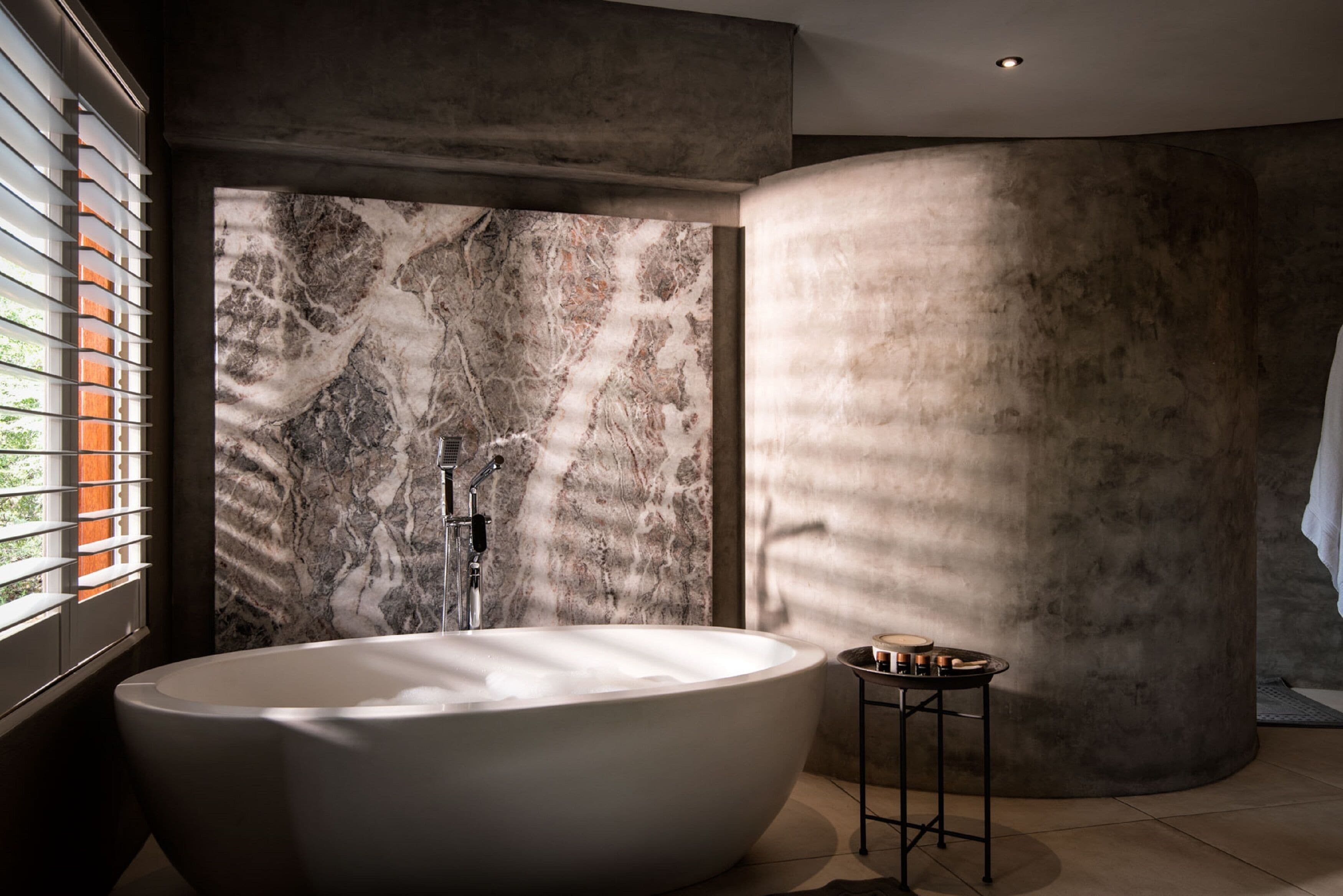 Un baño moderno con una bañera independiente, pared de mármol y una pequeña mesa con artículos de aseo.