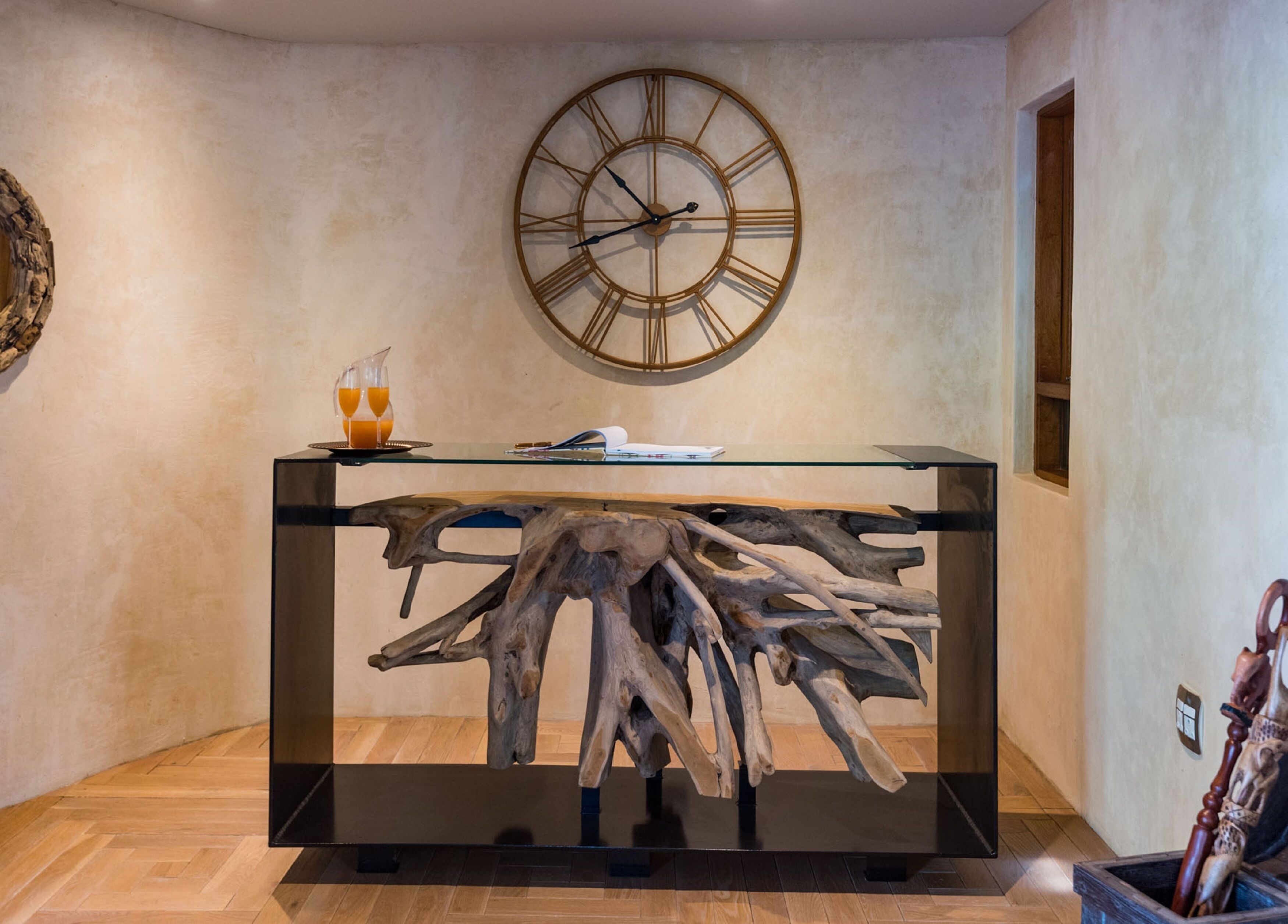 A modern interior with a wooden coffee table featuring a unique tree root design, a large wall clock, and a glass-top console table with a book and glasses.