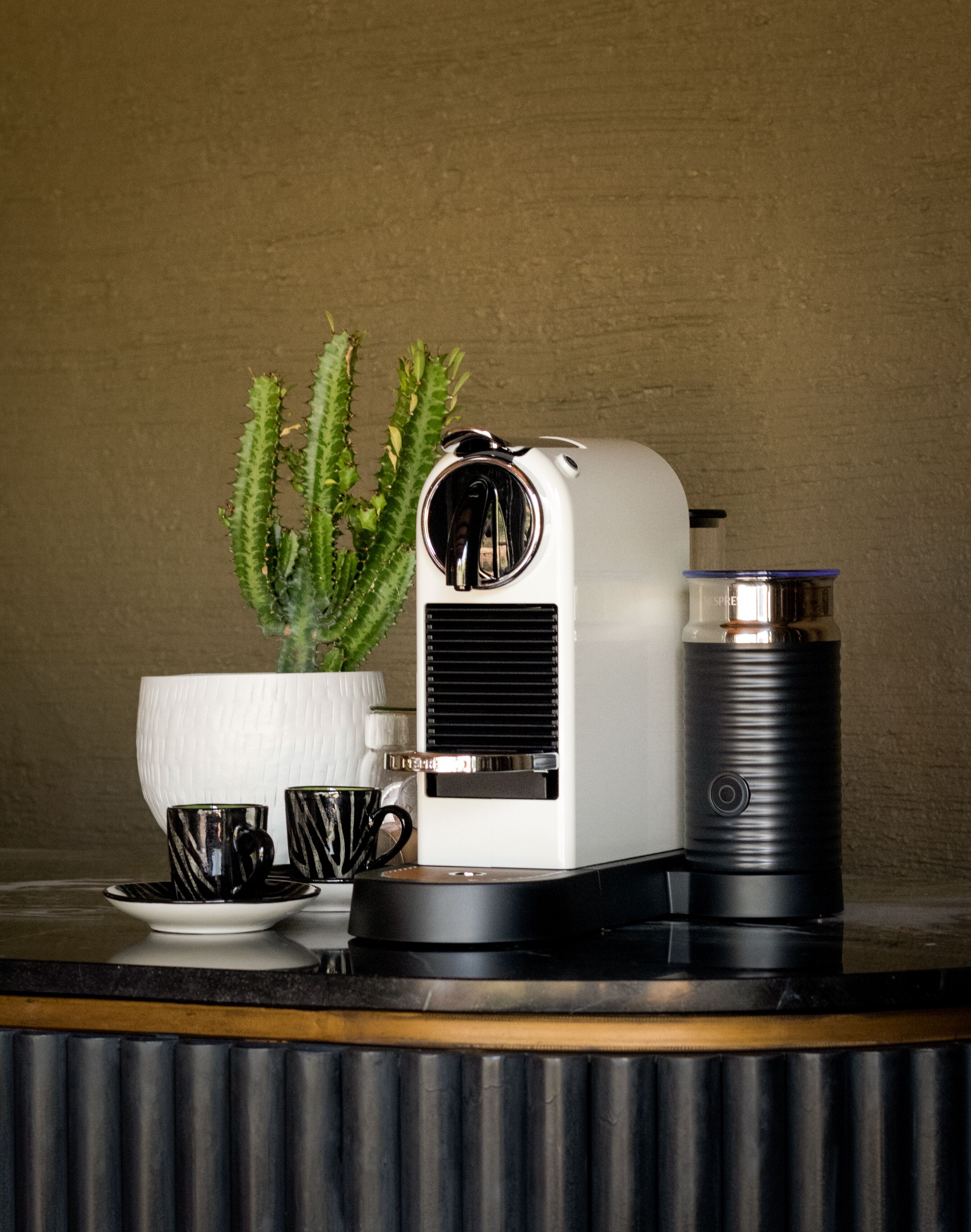 Uma cafeteira, uma garrafa térmica, um cacto e duas chávenas de café sobre uma mesa.