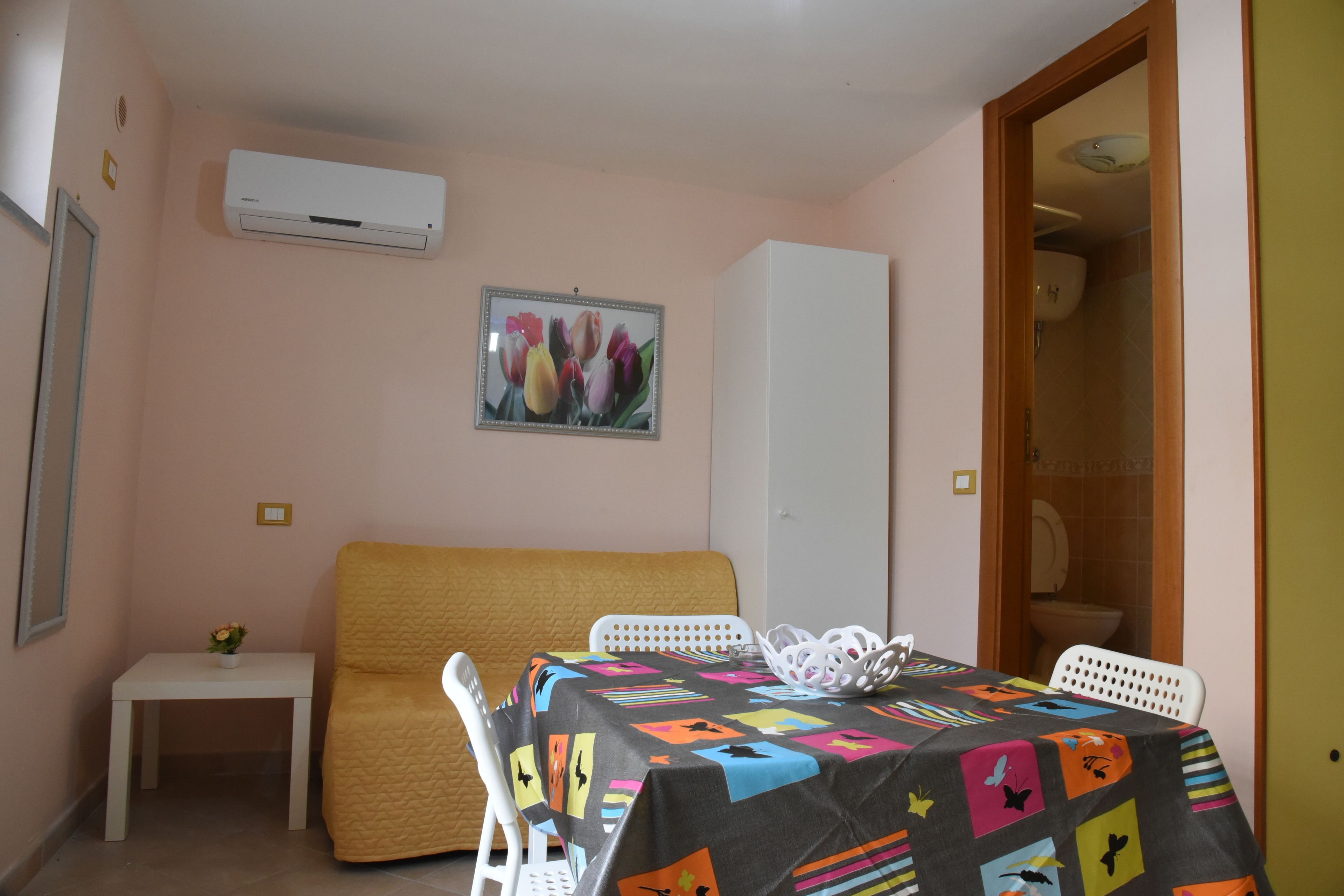A small room with a bed, a table with a colorful tablecloth, a white chair, a small white table with a flower, and an air conditioning unit on the wall.