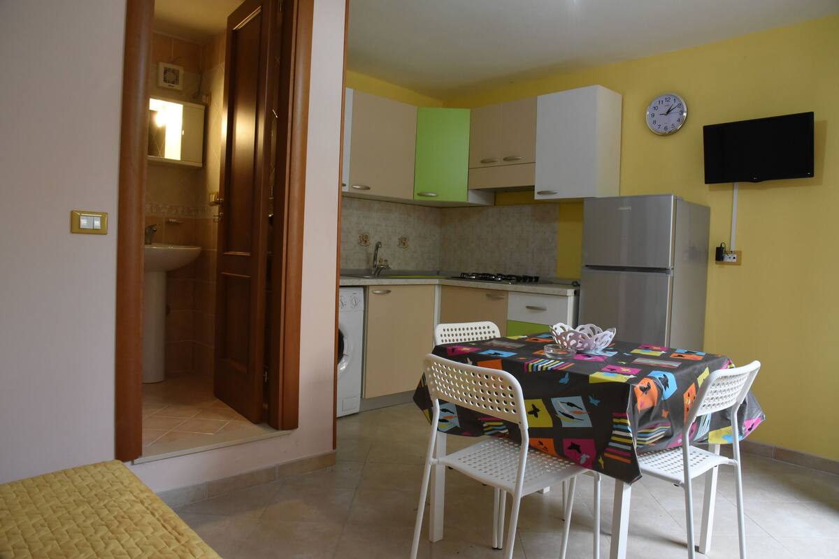 A compact kitchen with a dining area, including a table with chairs, a refrigerator, and a wall-mounted TV.