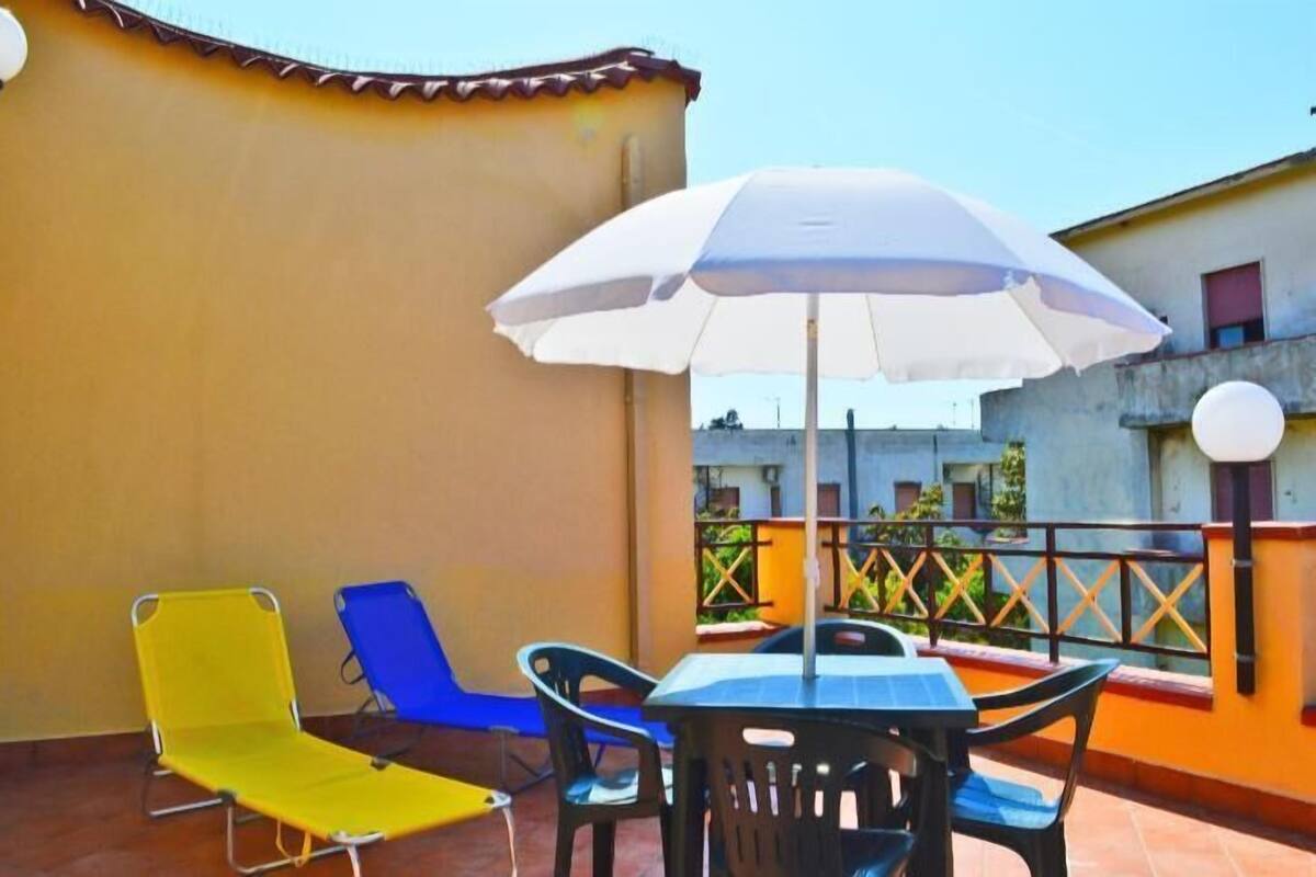 A terrace with a table and chairs, a white umbrella, and two colorful lounge chairs.