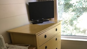 A room with a wooden desk, a yellow dresser with drawers, a television on top, and a window with a view of trees. - The Canberra Guest House (Wellingborough)