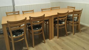A dining area with a rectangular wooden table surrounded by six wooden chairs with cushioned seats. - The Canberra Guest House (Wellingborough)