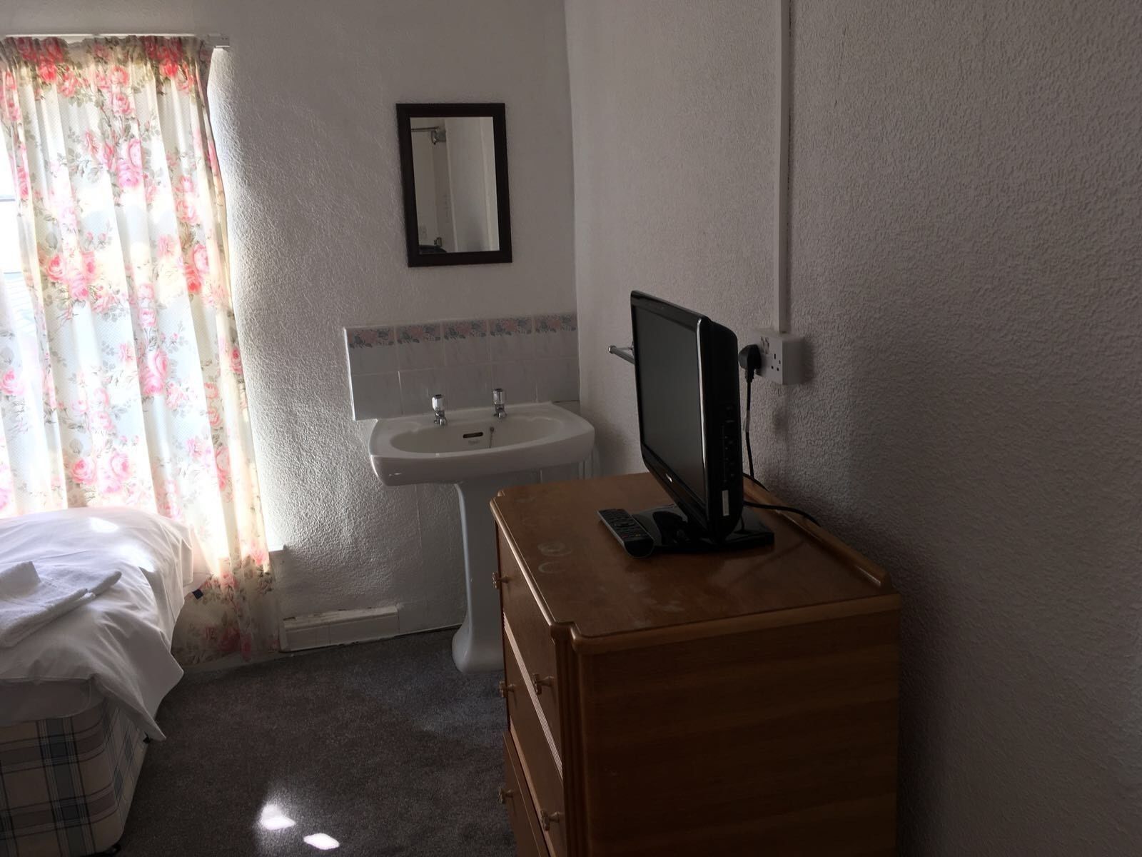 Un dormitorio con cama, cómoda de madera, televisor sobre un mueble, lavamanos y espejo en la pared.