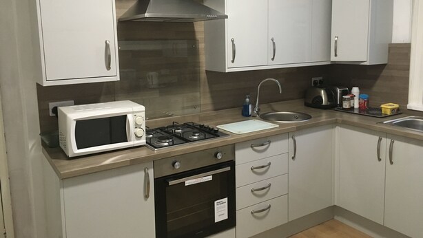 A modern kitchen with white cabinets, a stainless steel range hood, a microwave, a gas stove, a sink, and a dishwasher. - The Canberra Guest House (Wellingborough)