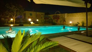 A nighttime swimming pool with three people in the water, surrounded by palm trees and a mural on the wall.