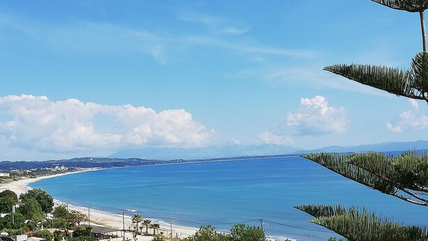 Vista costera con cielo despejado, playa y una cadena montañosa distante.