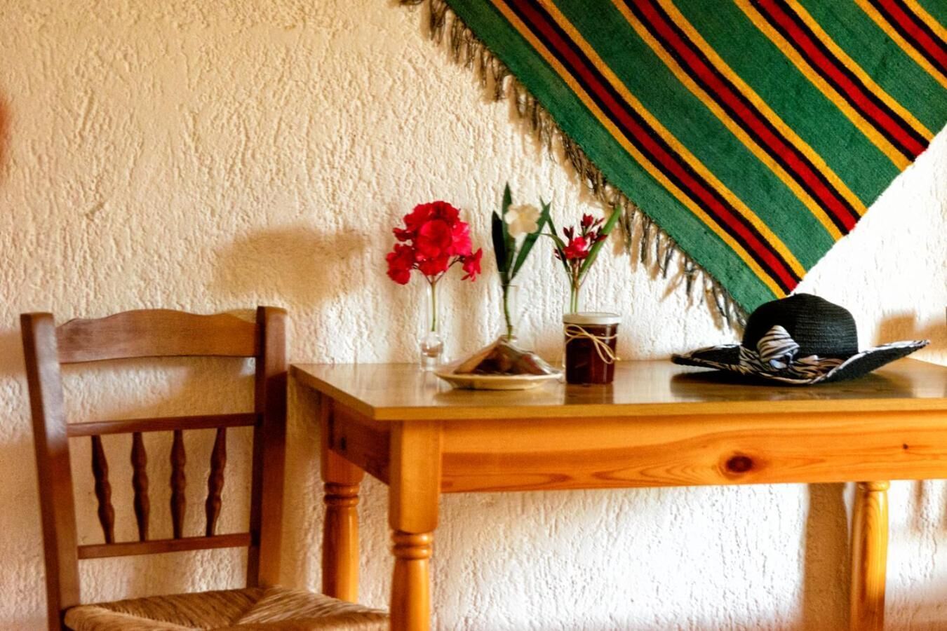 Een houten eettafel met een stoel, een vaas met rode bloemen, een pot en een zwarte hoed op tafel.