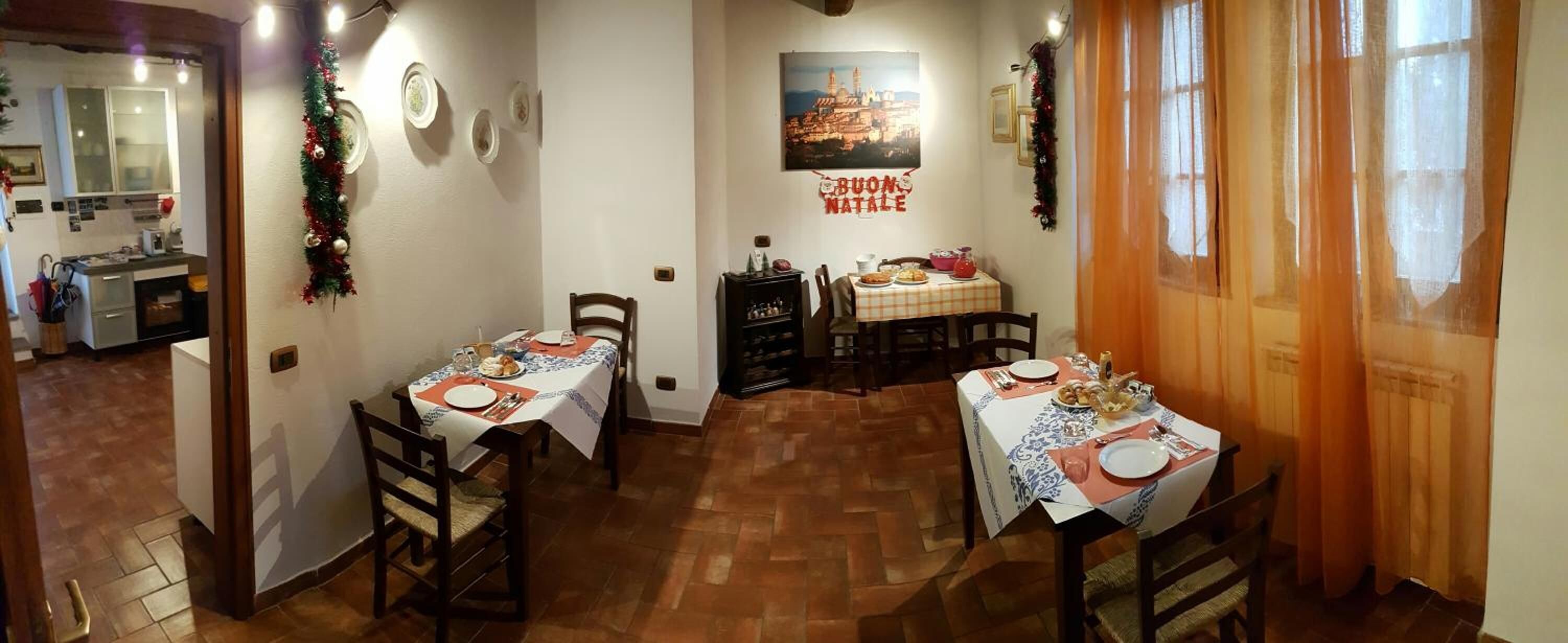Een eetruimte met twee tafels gedekt voor de maaltijden, een keuken op de achtergrond, en een 'Buon Natale' bord aan de muur.