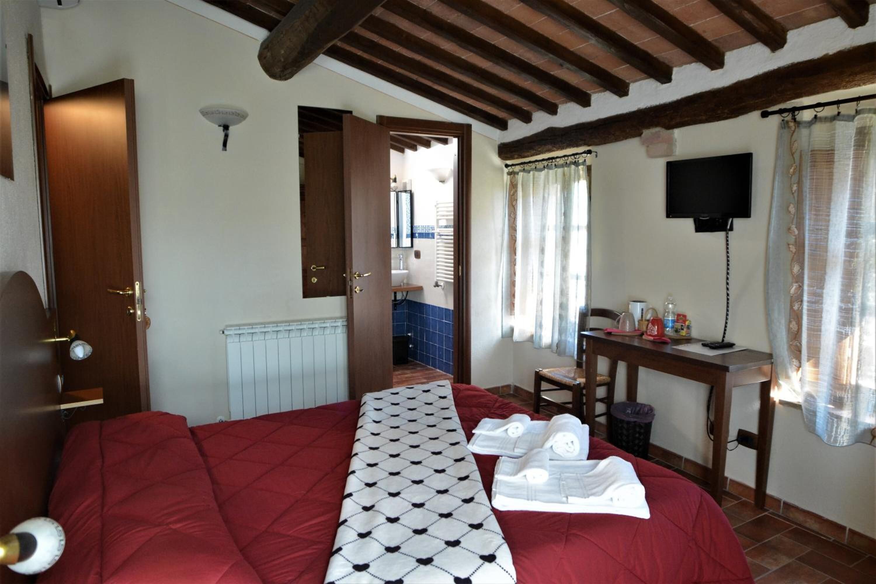 A bedroom with a large bed, a wooden door, a radiator, a desk with a chair, a TV, and a window with curtains.