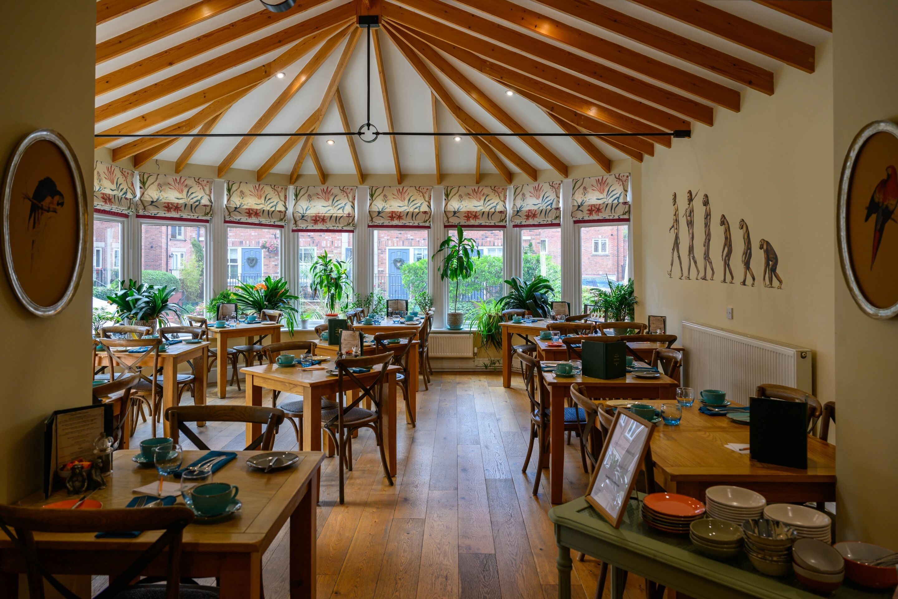 A dining area with wooden tables and chairs, large windows with plants, and a ceiling with wooden beams.