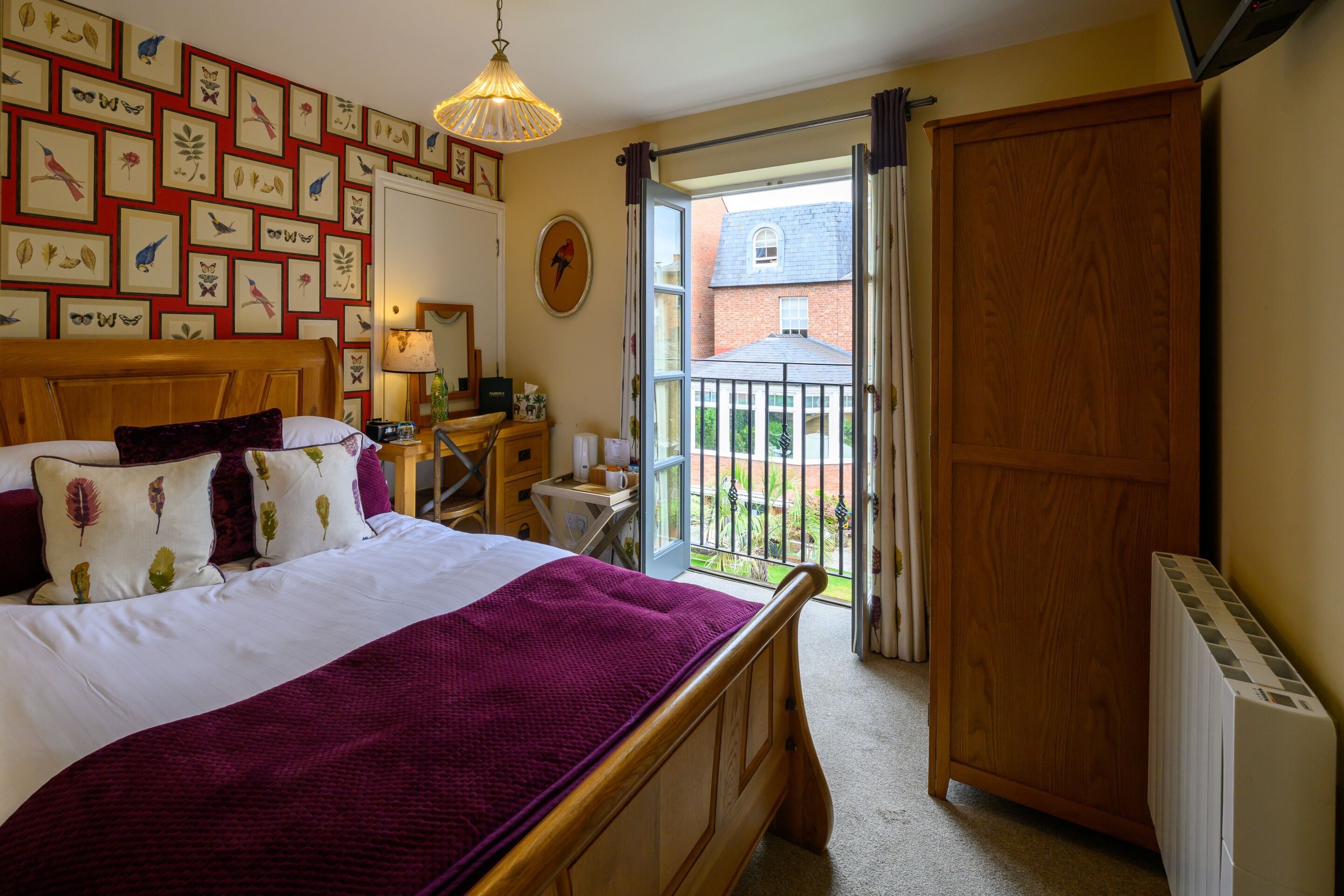 A bedroom with a large bed, a wooden headboard, a desk with a chair, a radiator, and a balcony with a view of a brick building.