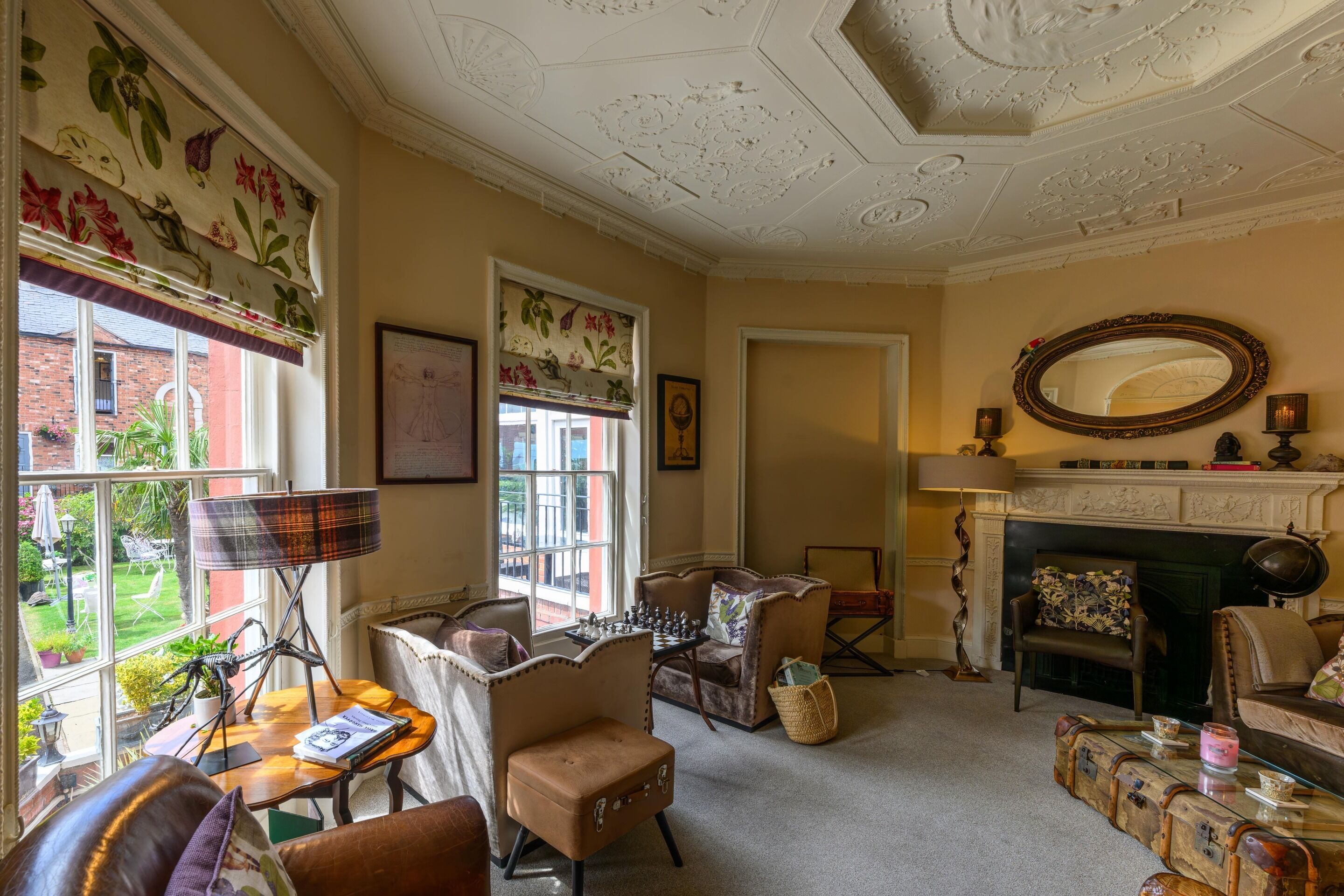 A cozy living room with a fireplace, comfortable seating, a chess table, and a view of the garden through large windows.