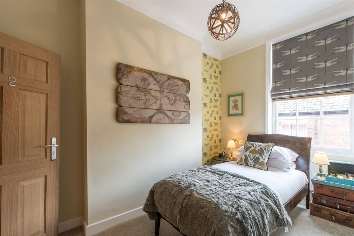 Een slaapkamer met een bed, een houten garderobe, een wereldkaart als kunstwerk, een raam met een geborduurd raamdecoratie en een nachtkastje met een lamp.