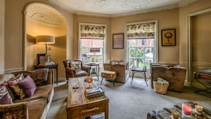 Een gezellige kamer met een bank, fauteuil en een salontafel. Er zijn twee ramen met bloemen gordijnen, een lamp en een wereldbol aan de muur.