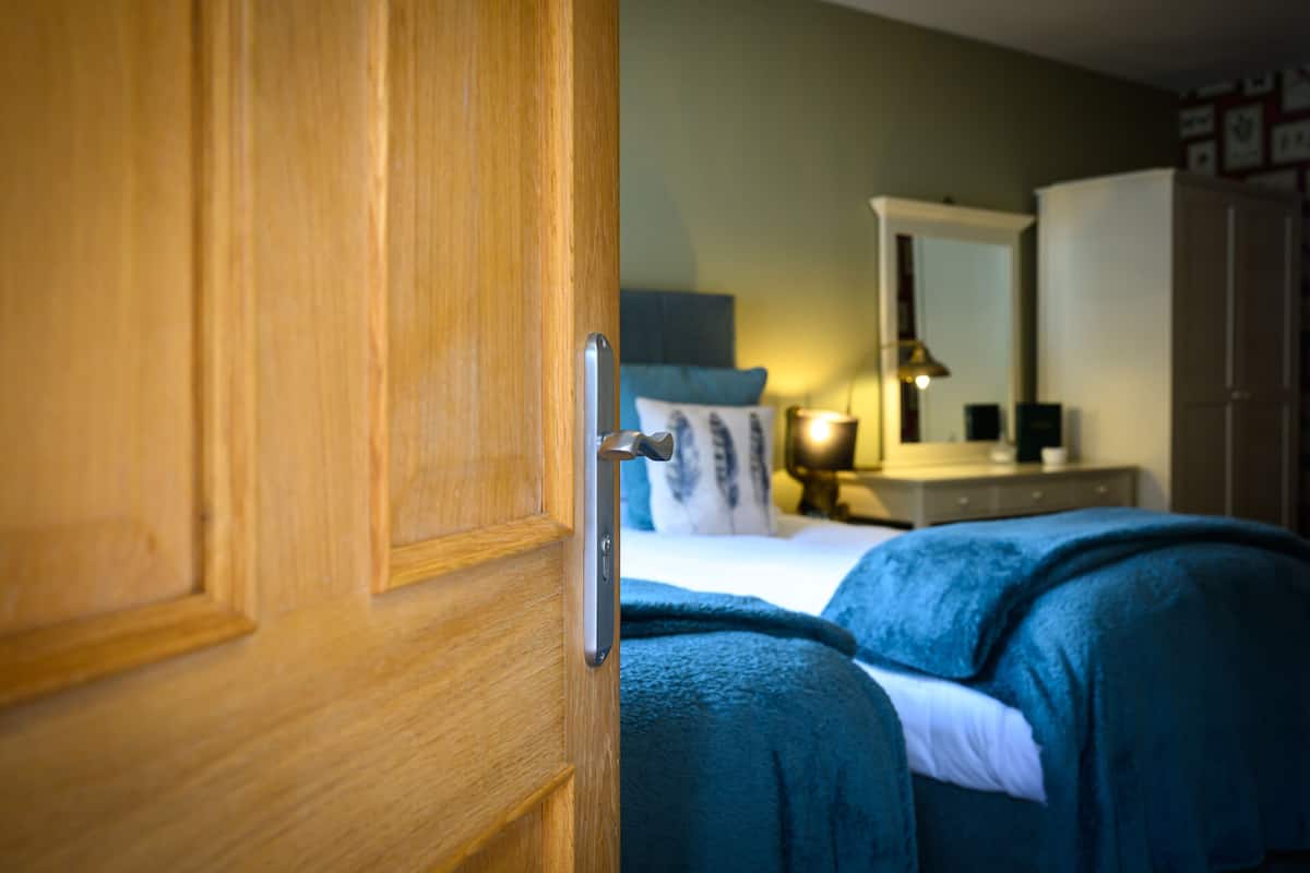 Een houten deur met een zilveren handvat, een bed met een blauwe deken, een nachtkastje met een lamp en een spiegel aan de muur.