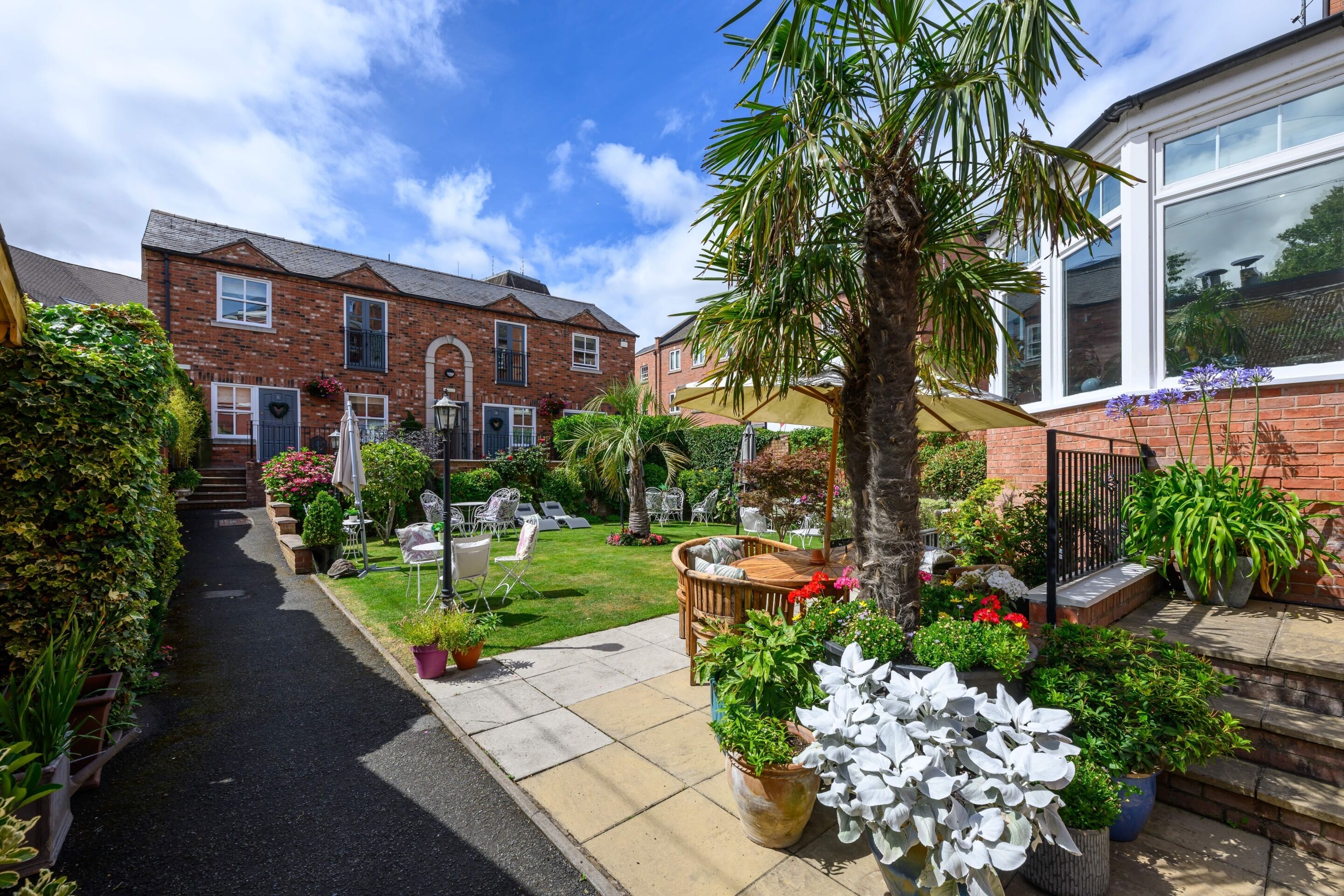 A residential garden with a paved pathway, outdoor seating, and various potted plants.