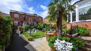 A residential garden with a paved pathway, outdoor seating, and various potted plants.
