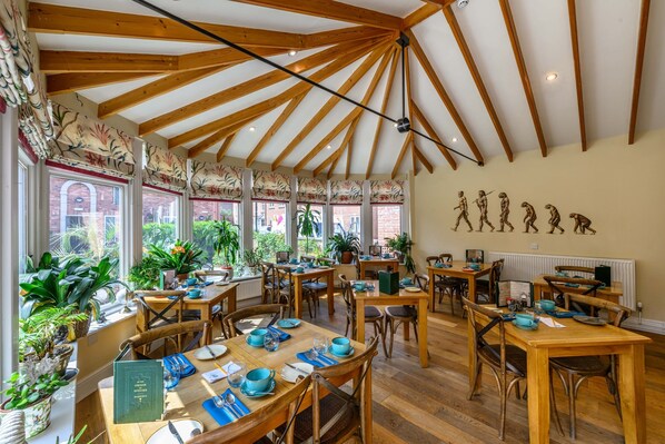 Un espace repas avec des tables et des chaises en bois, de grandes fenêtres avec des rideaux et un mur orné d’une fresque représentant un dinosaure.