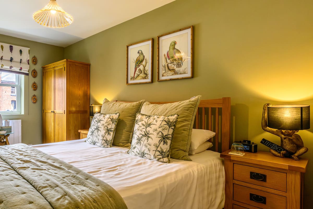 Een net opgemaakt bed met bloemenkussens, een houten nachtkastje met lamp en afstandsbediening, en twee ingelijste vogelafbeeldingen aan de muur.