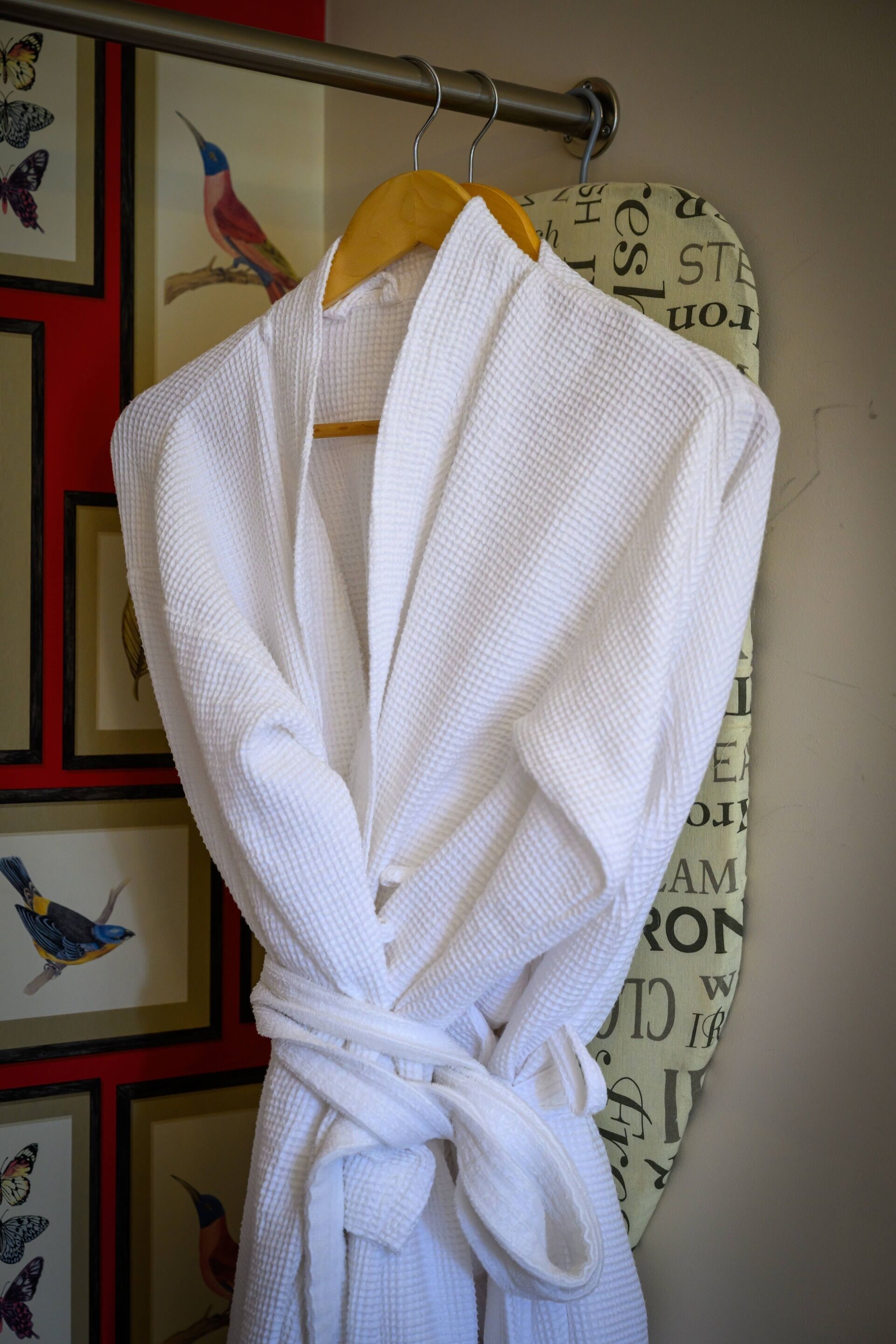 A white bathrobe hanging on a wooden hanger against a wall with framed bird illustrations.
