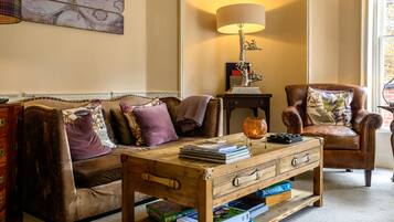 Uma sala de estar acolhedora com um sofá de couro marrom, uma mesa de centro de madeira com livros e jogos de Scrabble, um abajur e um mapa na parede.