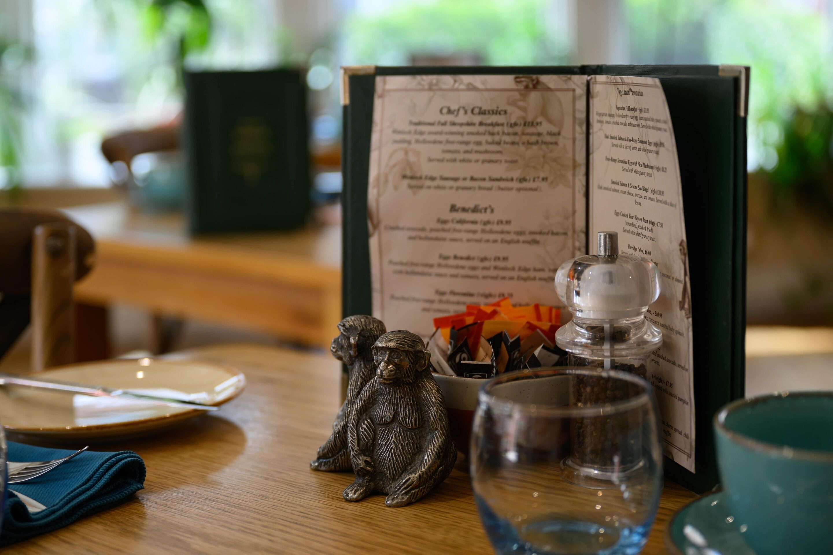 A menu with various dishes listed, a small monkey figurine, a glass, and a bottle on a wooden table.