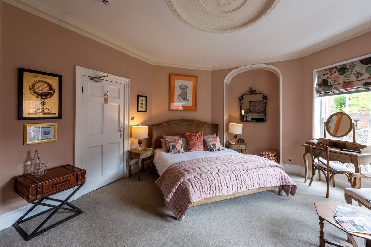 Een slaapkamer met een groot bed, nachtkastjes, een bureau en een stoel. Er is een deur, een raam met gordijn en wanddecoraties.