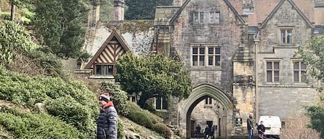 Un edificio histórico de piedra con un arco de entrada destacado, rodeado de exuberante vegetación y un camino pavimentado.