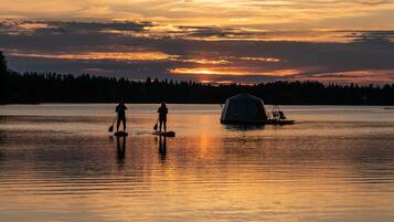 Un lago sereno al atardecer, con dos personas haciendo paddleboard y una estructura cubierta sobre el agua.