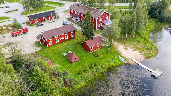 A red two-story house with a dock by a lake, surrounded by greenery and a gravel driveway.