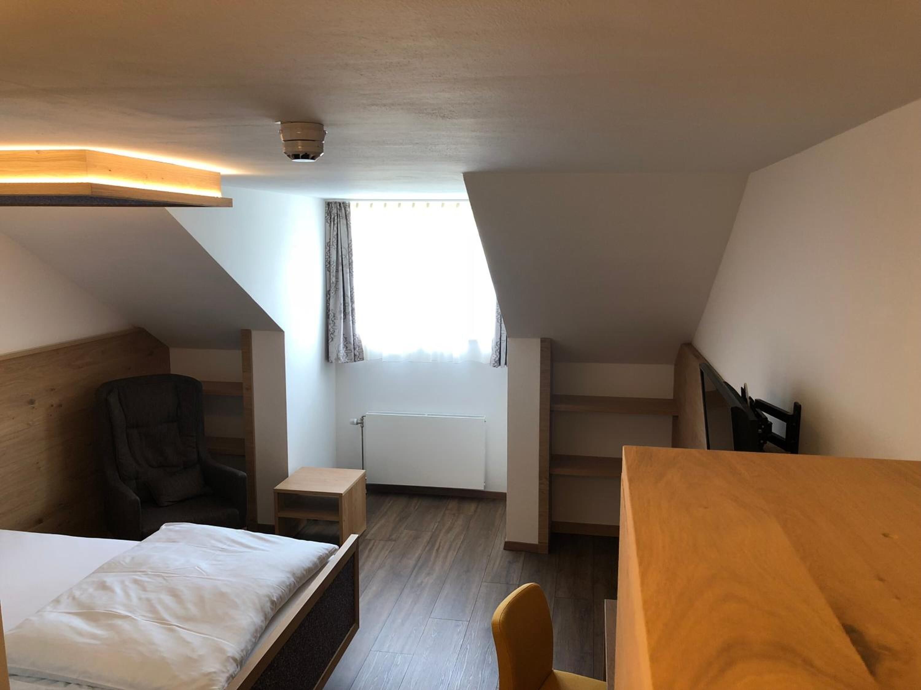 A compact hotel room with a bed, a desk, a chair, a radiator, and a window with curtains.