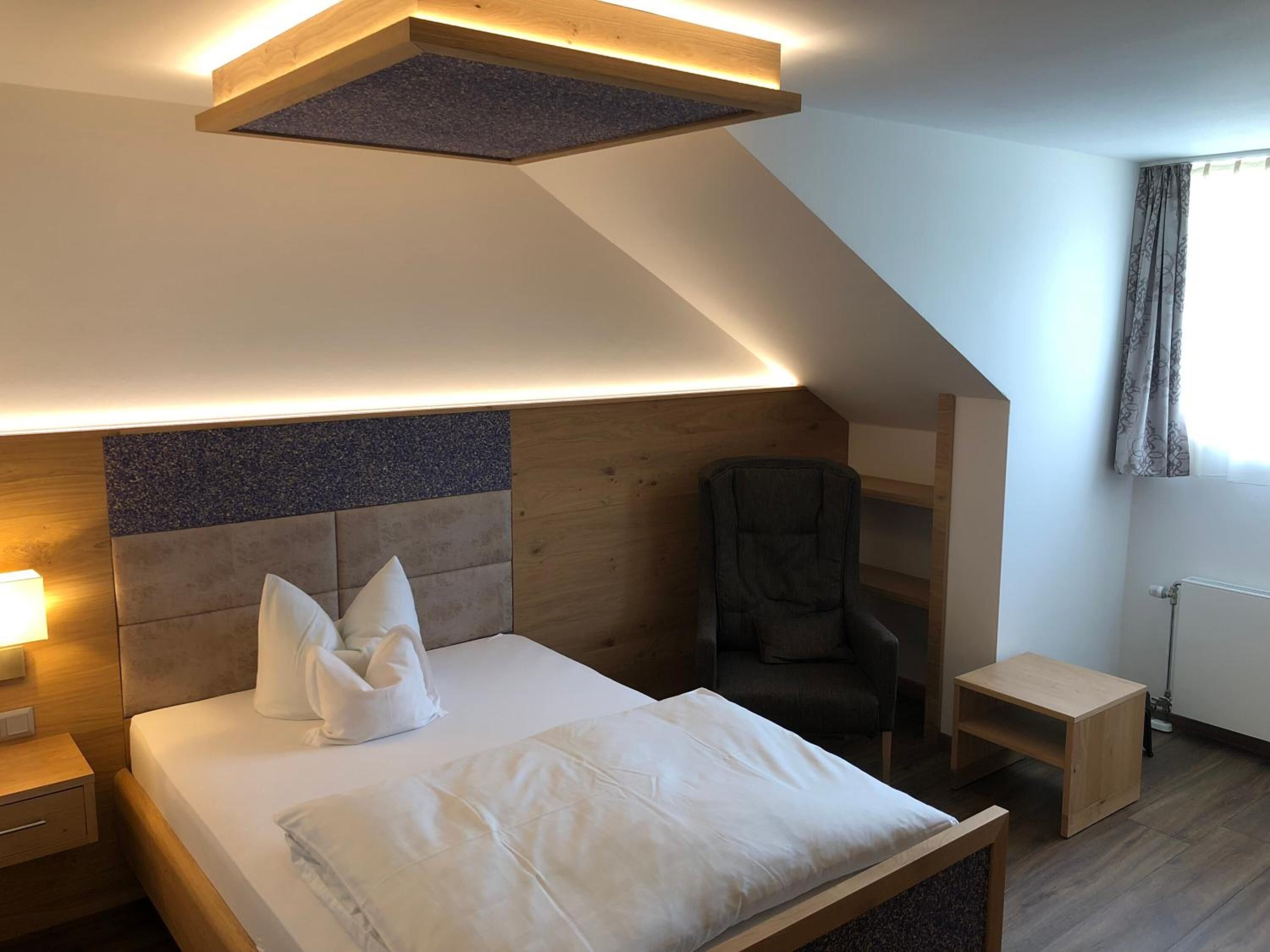 Quarto moderno com teto inclinado, cama com roupa de cama branca, cabeceira de madeira, uma cadeira e uma pequena mesa de madeira.