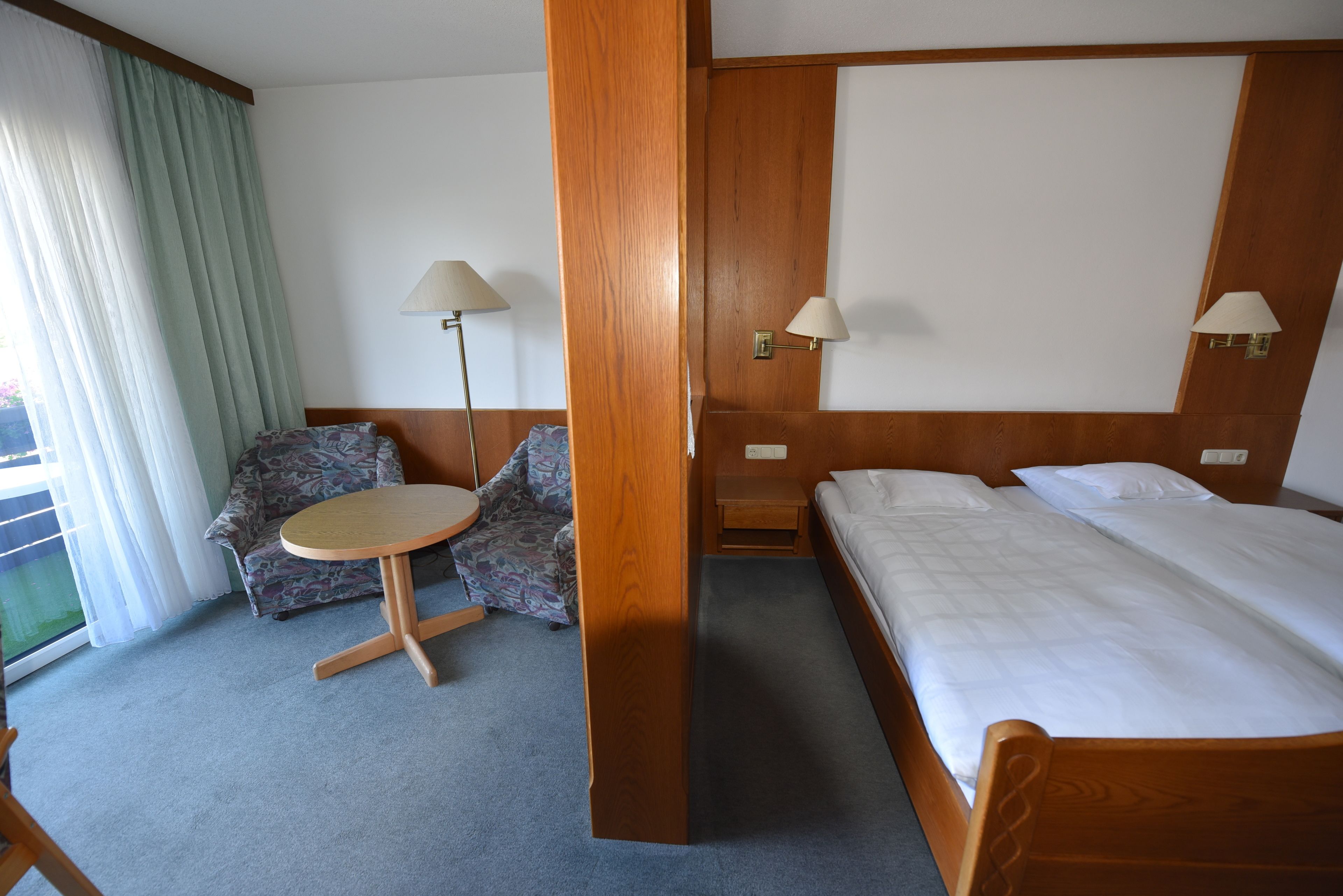 A hotel room with a bed, a chair, a small table, and two wall-mounted lamps.