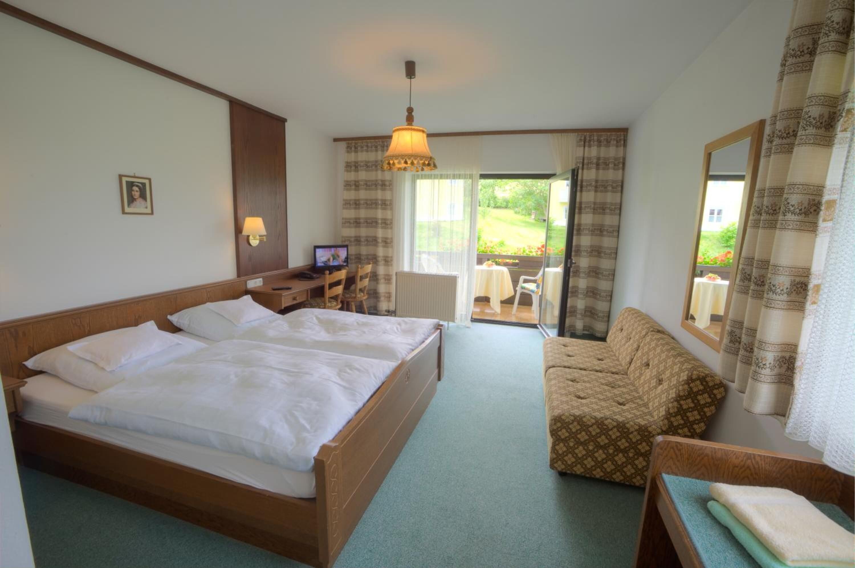 A hotel room with a large bed, a sofa, a small table, a TV, and a view of the garden through the sliding doors.