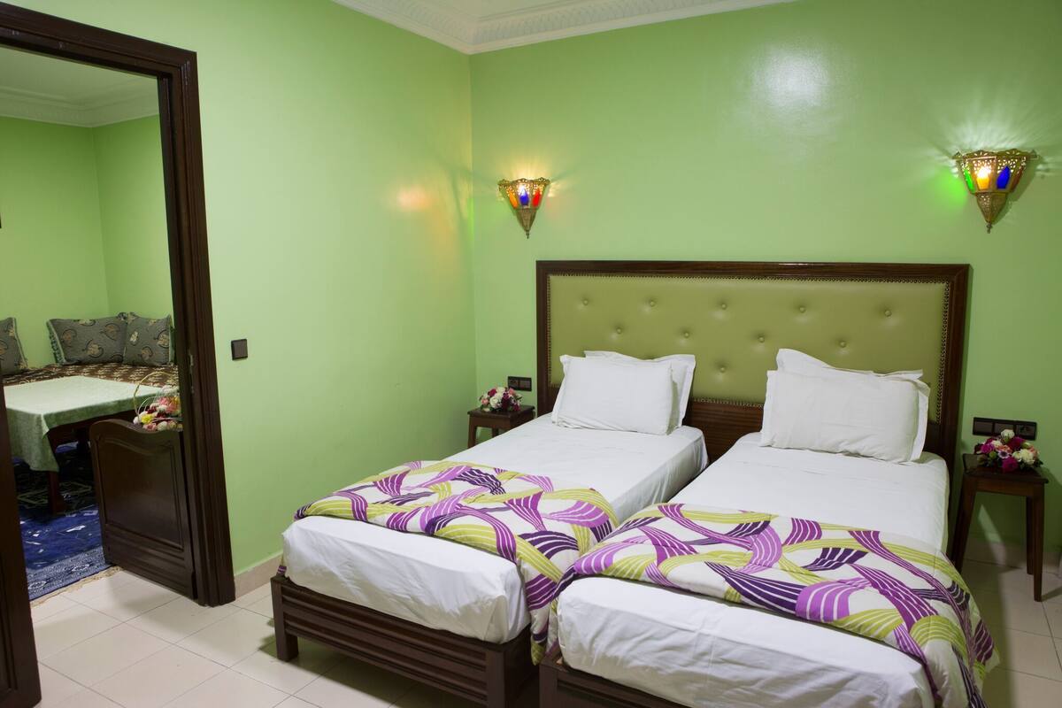A hotel room with two beds, a dining table, and two wall-mounted lamps.