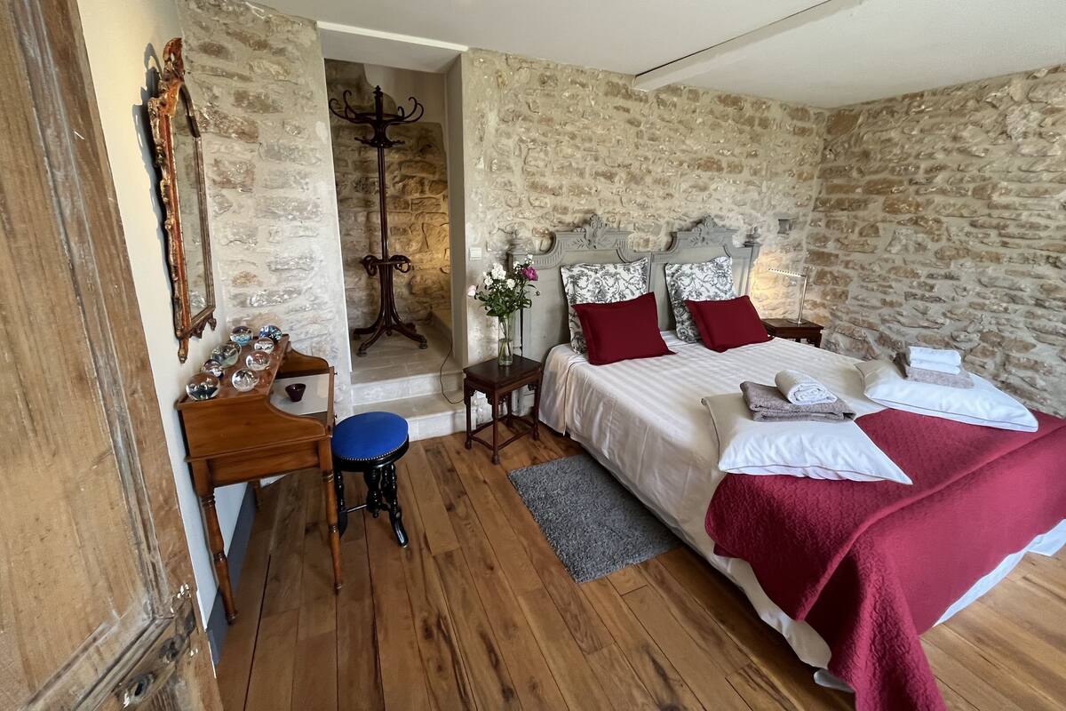 A bedroom with a stone wall, a wooden bed, a desk with a chair, and a mirror.