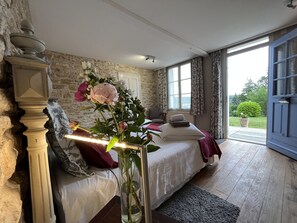 A bedroom with a stone wall, a bed with white bedding, a vase with flowers, and a view of the outdoors through a large window. - Chenevieres d'en Haut (Grimault)