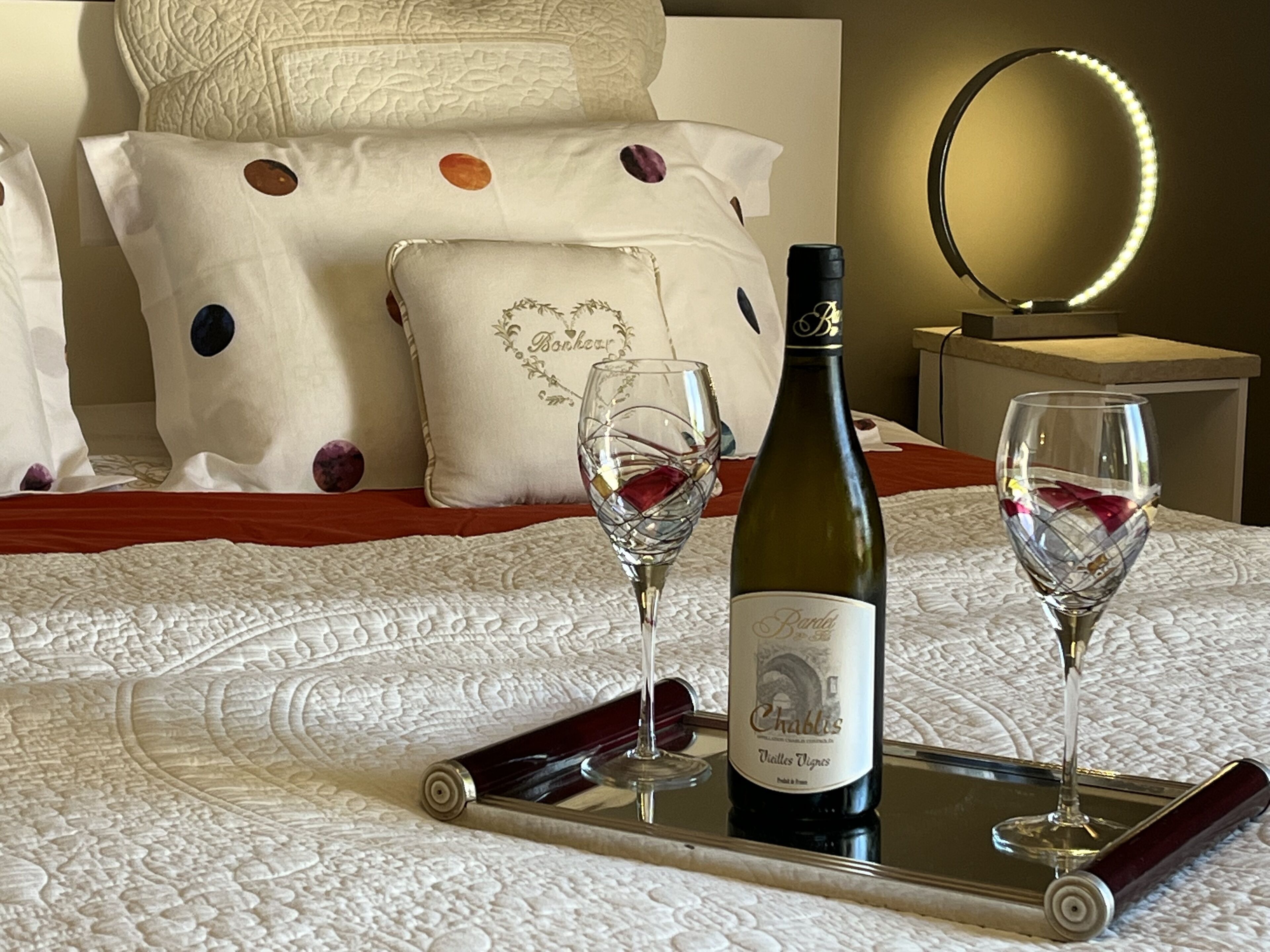 Un lit recouvert d’une couette blanche, une bouteille de vin et deux verres à vin posés sur un plateau.