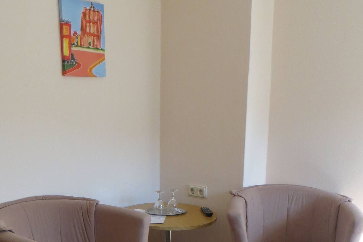 A hotel room with two armchairs, a small round table with a remote control, a glass set, and a painting of a famous building.
