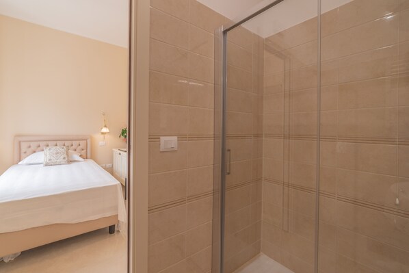 A bedroom with a bed, a nightstand, a lamp, and a glass-enclosed shower area. - Hotel Casa Adele (Taormina)