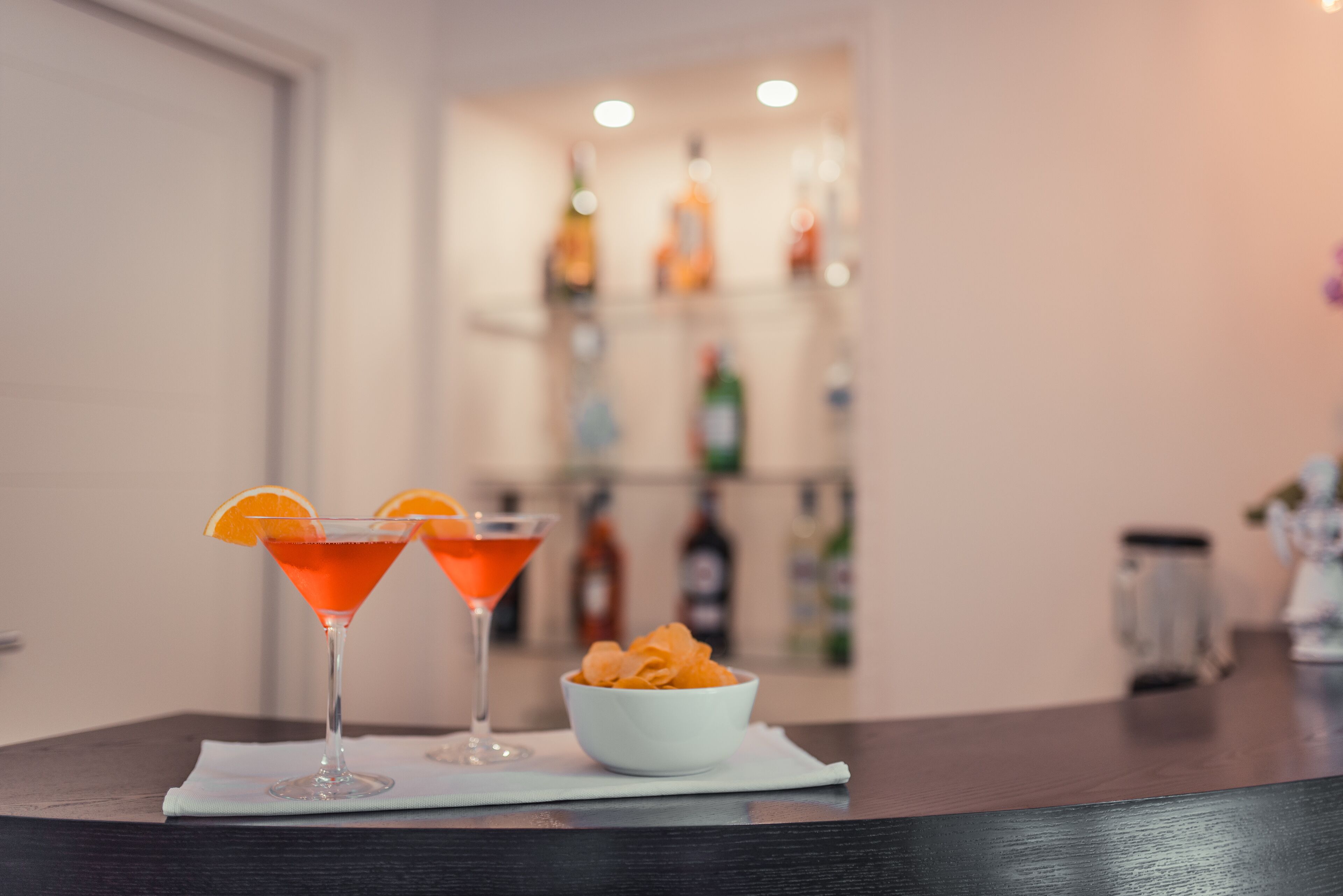 Two martini glasses with orange garnishes on a table, a bowl of orange snacks, and a blurred background with shelves of bottles.