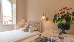 Habitación de hotel con cama, lámparas de noche, mesita de noche, un florero con planta y ventana con puerta abierta.