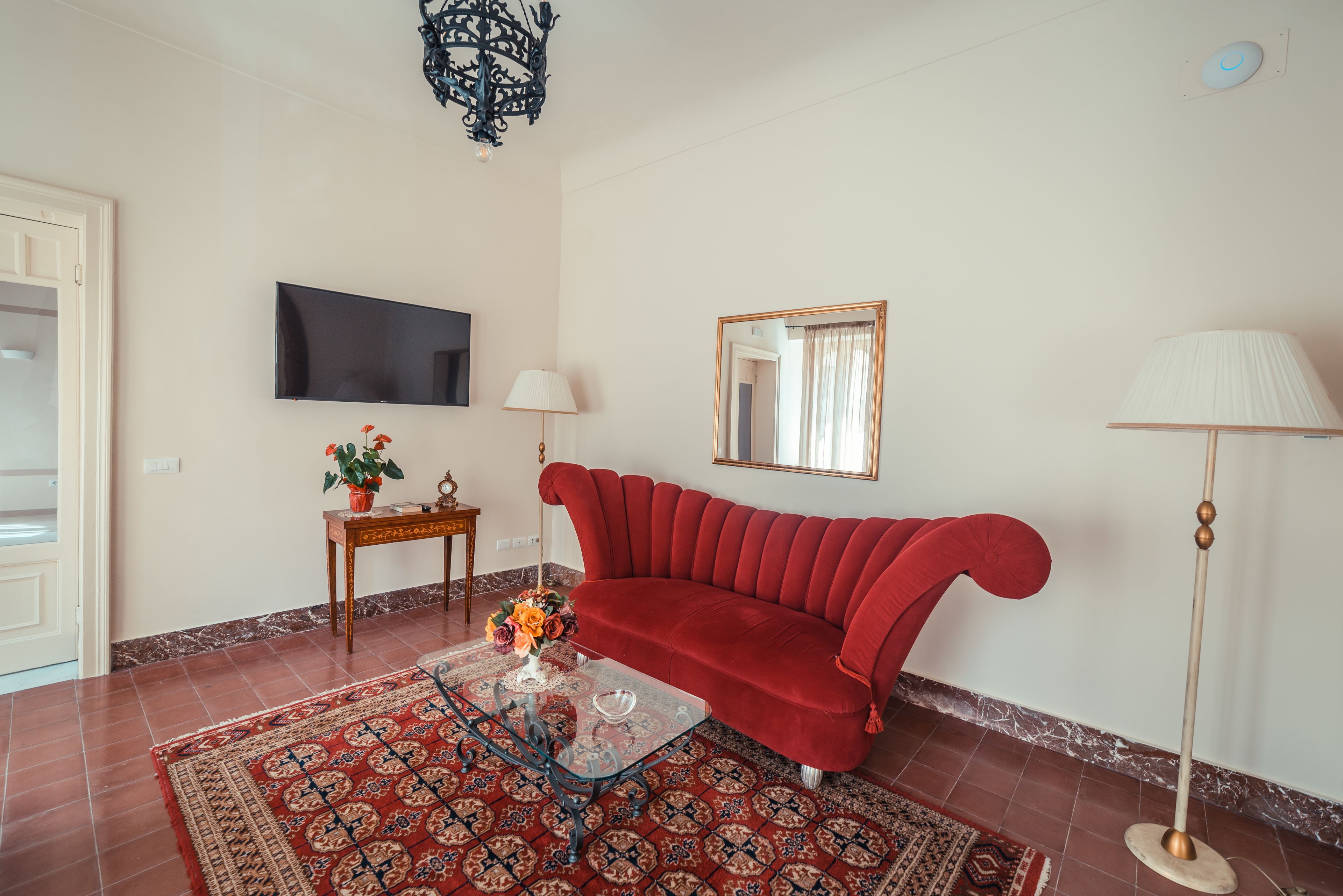 Een woonkamer met een rode bank, een glazen salontafel, een kamerkleed met dessin, een televisie, een lamp en een spiegel.