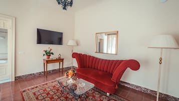 Een woonkamer met een rode bank, een glazen salontafel, een kamerkleed met dessin, een televisie, een lamp en een spiegel.